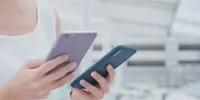 Pessoa segurando dois smartphones, um com capa roxa e outro azul, em ambiente moderno e iluminado, representando comparação ou uso simultâneo de dispositivos móveis.