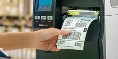 Mão segurando etiqueta recém‑impressa em impressora industrial Zebra ZT600, com códigos de barras e informações de rastreamento, representando impressão de etiquetas para logística e controle de estoque.